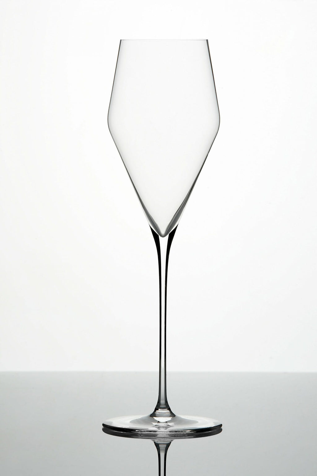 Ein hohes, leeres, klares Weinglas mit langem Stiel steht auf einer reflektierenden Oberfläche. Das Glas hat ein markantes, eckiges Kelchdesign, das sich nach oben hin verjüngt, was ihm ein modernes und elegantes Aussehen verleiht. Der Hintergrund ist schlicht und sanft beleuchtet.