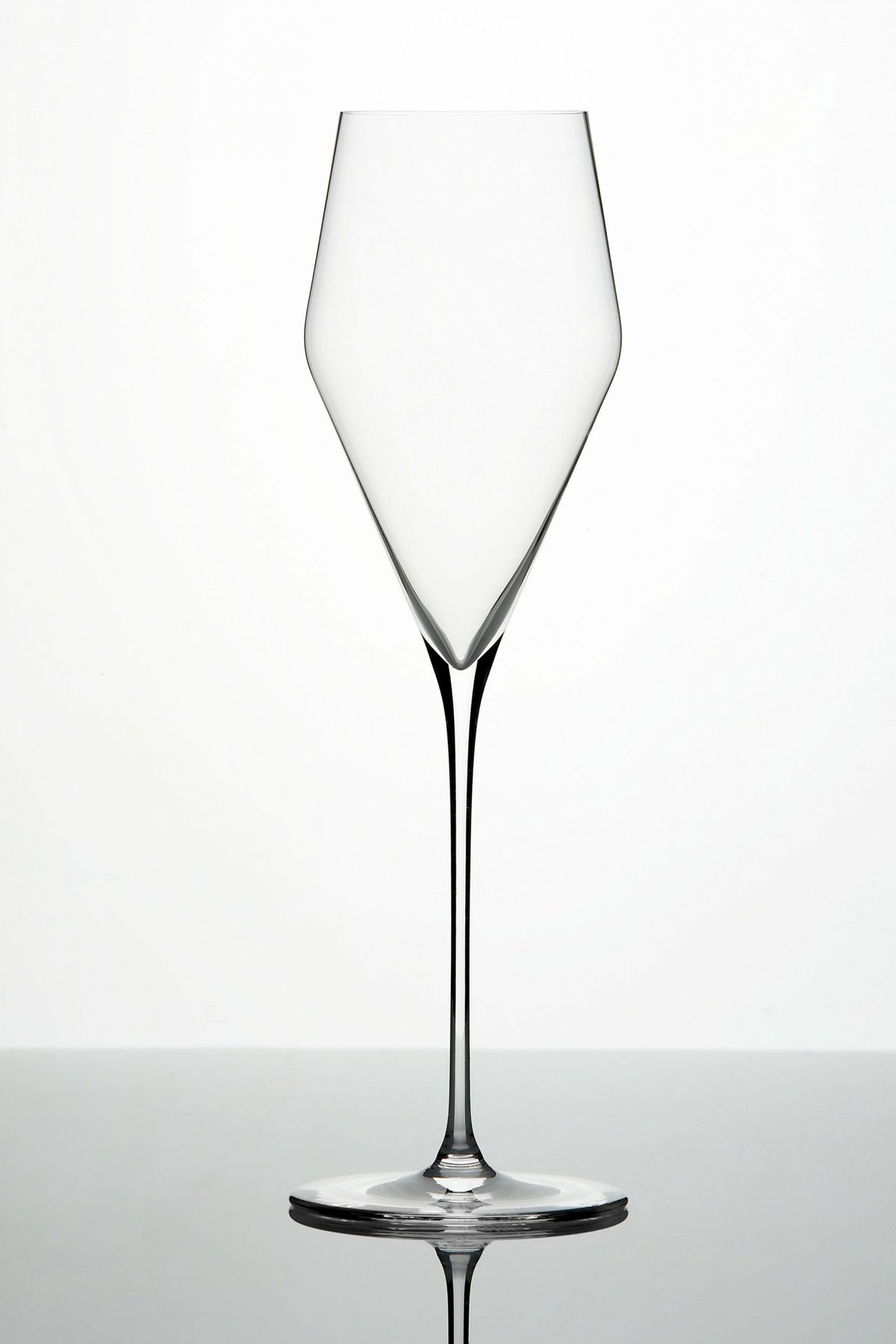 Ein hohes, leeres, klares Weinglas mit langem Stiel steht auf einer reflektierenden Oberfläche. Das Glas hat ein markantes, eckiges Kelchdesign, das sich nach oben hin verjüngt, was ihm ein modernes und elegantes Aussehen verleiht. Der Hintergrund ist schlicht und sanft beleuchtet.