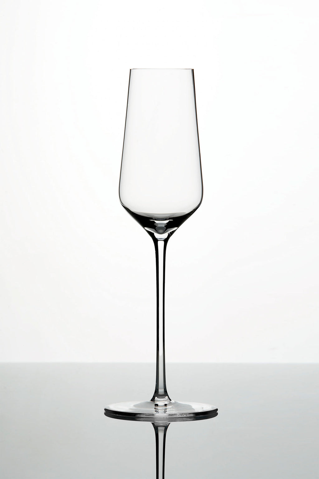 Ein klares, leeres Weinglas mit langem Stiel und leicht konischer Schale steht vor einem schlichten weißen Hintergrund. Das Glas ruht auf einer reflektierenden Oberfläche und spiegelt sein elegantes und minimalistisches Design wider.