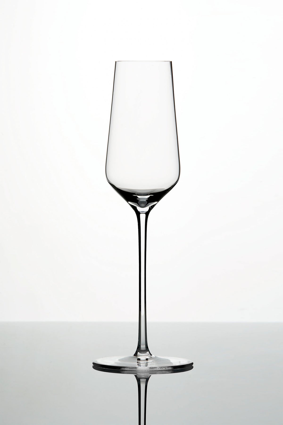 Ein klares, leeres Weinglas mit langem Stiel und leicht konischer Schale steht vor einem schlichten weißen Hintergrund. Das Glas ruht auf einer reflektierenden Oberfläche und spiegelt sein elegantes und minimalistisches Design wider.