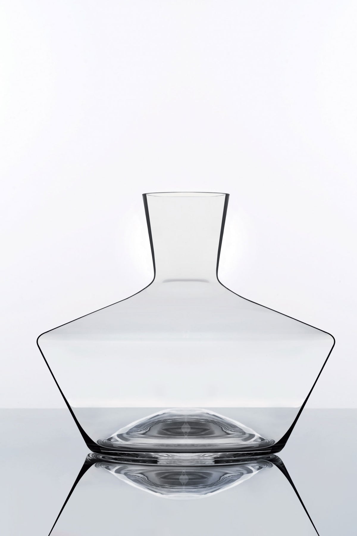 Eine minimalistische Karaffe aus klarem Glas mit breitem Boden und schmalem Hals steht vor einem schlichten weißen Hintergrund. Die Reflexionen der darunterliegenden verspiegelten Oberfläche erzeugen einen symmetrischen Effekt und betonen die klaren Linien der Karaffe.