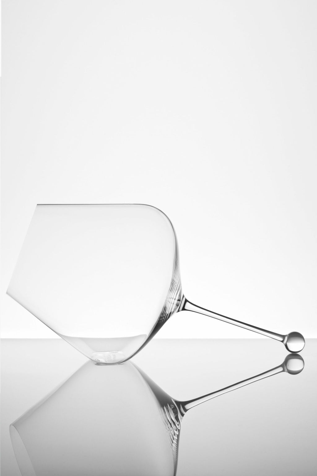 Ein minimalistisches Foto eines klaren, leeren Weinglases, das auf der Seite liegt, vor einem weißen Hintergrund. Seine Spiegelungen sind auf der glatten Oberfläche darunter sichtbar und erzeugen eine künstlerische und symmetrische Komposition.