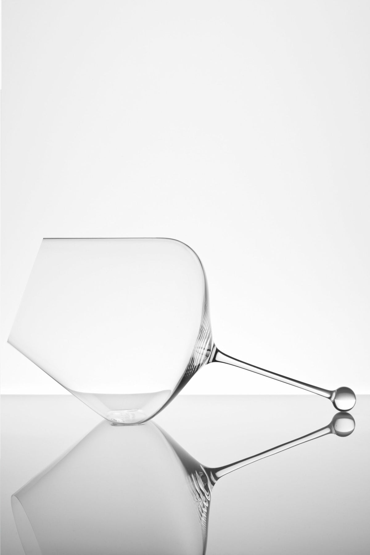 Ein minimalistisches Foto eines klaren, leeren Weinglases, das auf der Seite liegt, vor einem weißen Hintergrund. Seine Spiegelungen sind auf der glatten Oberfläche darunter sichtbar und erzeugen eine künstlerische und symmetrische Komposition.