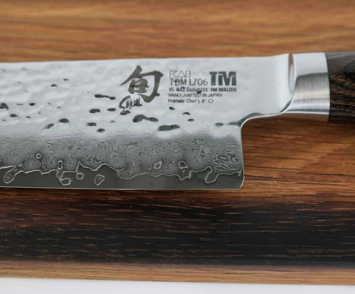 Eine Nahaufnahme des KAI Shun Premier Tim Mälzer TDM-1706, ein ultrascharfes japanisches Messer aus Damaststahl mit detaillierten Ätzungen auf der 20 cm langen Klinge und einem dunklen Holzgriff. Die Klinge weist Kanji-Zeichen und englischen Text auf, was darauf hinweist, dass sie in Japan aus VG-MAX-Superstahl handgefertigt wurde. Im Hintergrund ist ein handgefertigtes Küchenbrett aus der Pfalz zu sehen, das 30 x 18 cm misst und aus Weinfassholz gefertigt ist.