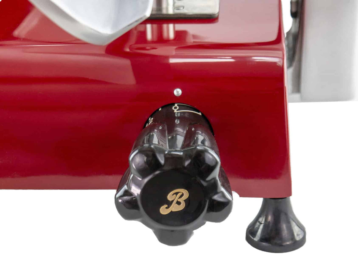 Eine Nahaufnahme einer Berkel Red Line 300 Rot, Original Profi-Aufschnittmaschine zeigt eine rote Maschine mit einem schwarzen Einstellknopf im Vordergrund. Der Knopf mit einem „B“-Logo hat an seiner Basis nummerierte Markierungen zum Einstellen von Einstellungen. Über dem Knopf mit 300 mm Messerdurchmesser ist teilweise ein graues Metallteil sichtbar.